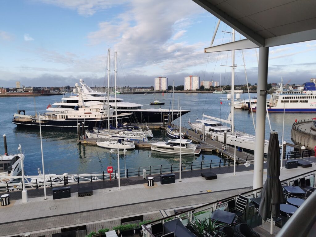 Luxusyacht und Segelboote am Kai, daneben eine Fähre; dahinter Stadtpanorama unter teils bewölktem Himmel.