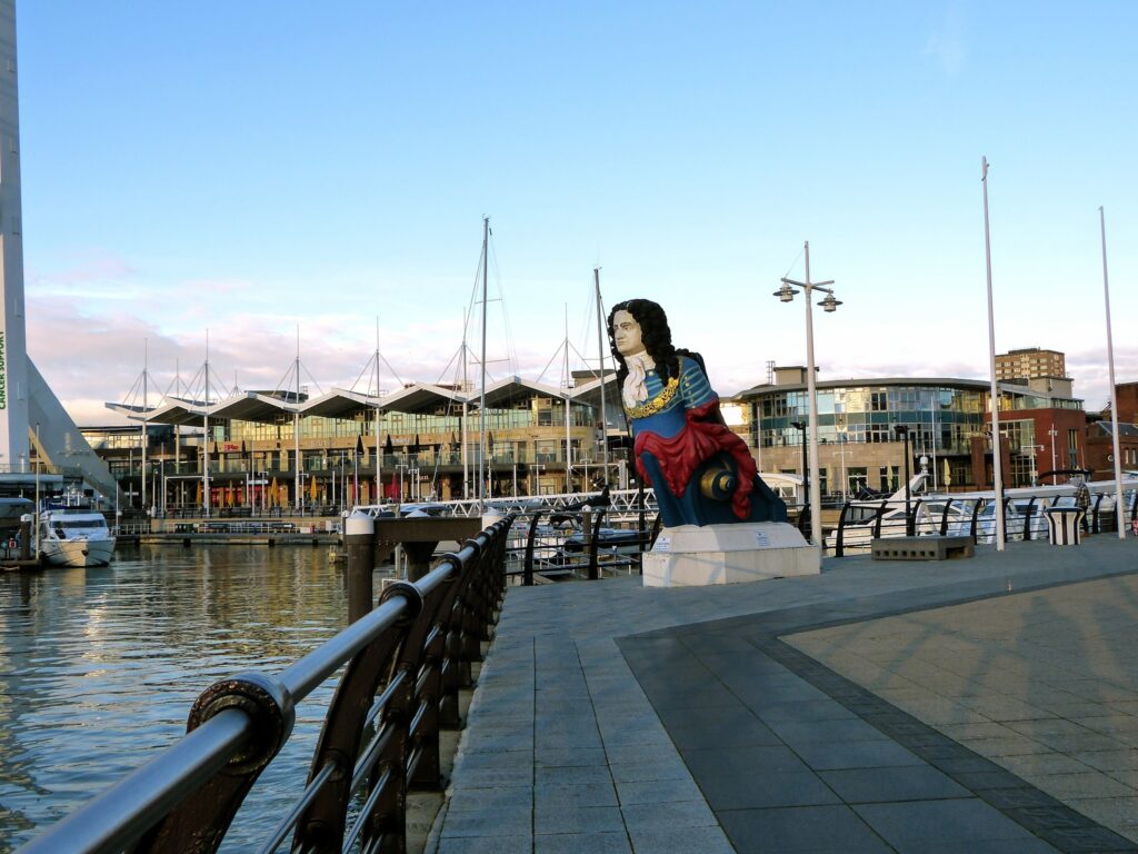 Große bunte Galionsfigur eines Admirals am Hafensteg, dahinter Yachten und moderne Glasgebäude im Abendlicht.
