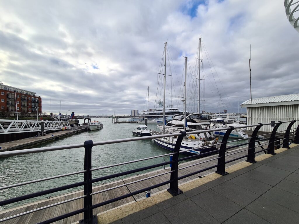 Yachthafen mit Segelbooten am Steg, grauer Himmel, Promenade mit Geländer im Vordergrund