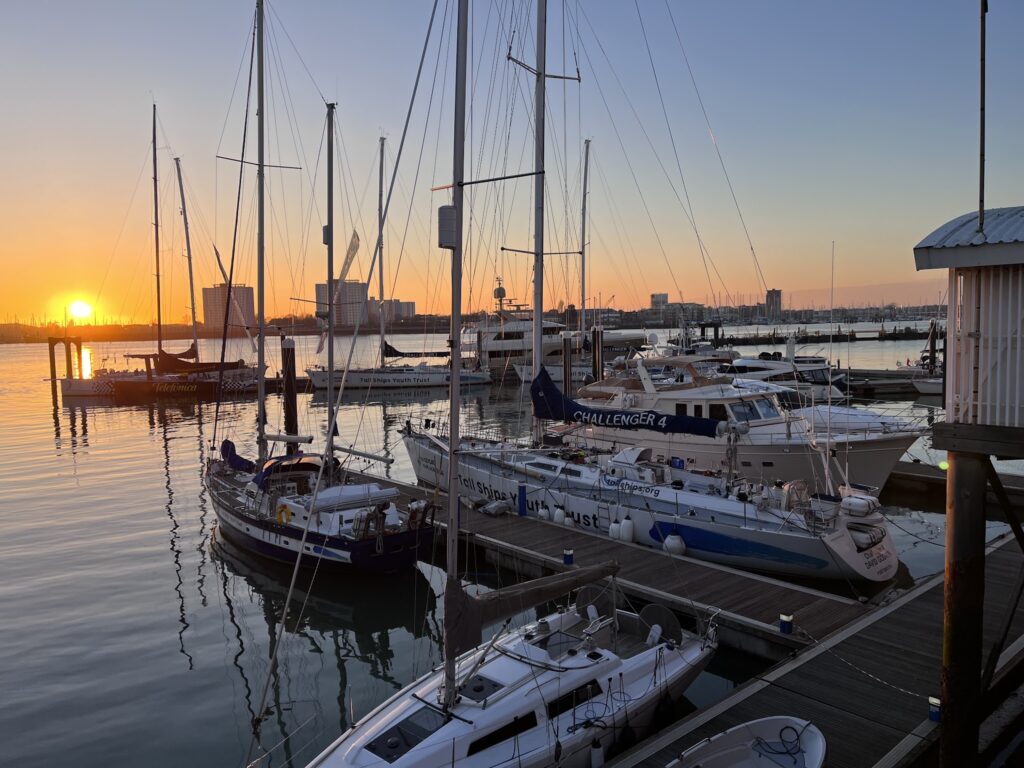 Sonnenuntergang über einem Yachthafen; stilles Wasser, Segelboote und hohe Masten vor orangeblauem Himmel