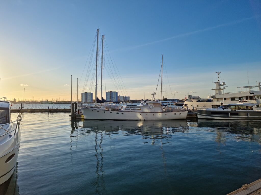 Yachten und Segelboote liegen im ruhigen Hafen; Abendlicht färbt Himmel und spiegelndes Wasser warm.