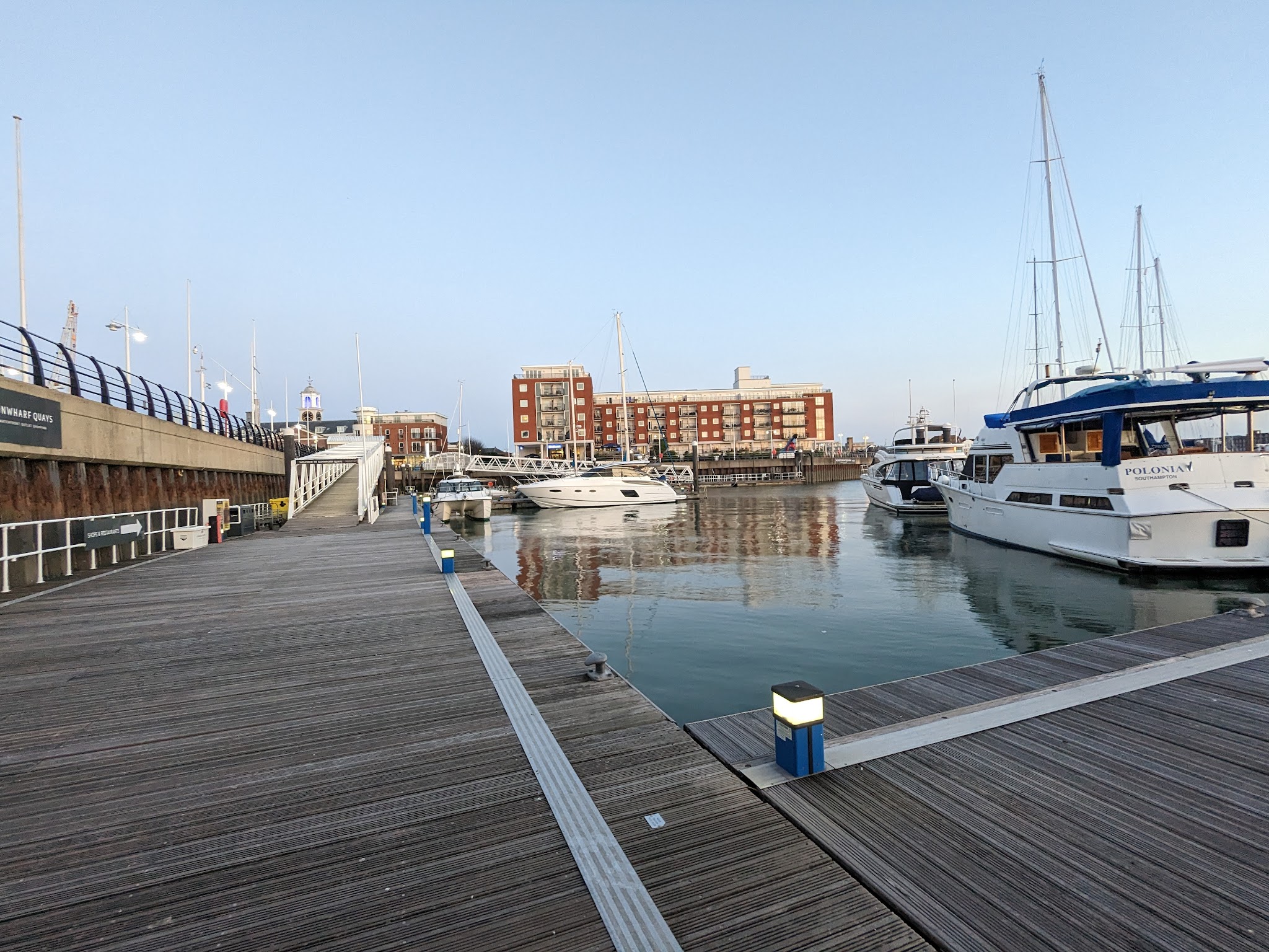 Holzsteg an ruhigem Hafen; mehrere weiße Yachten liegen vor Backstein-Wohnhäusern im sanften Abendlicht.