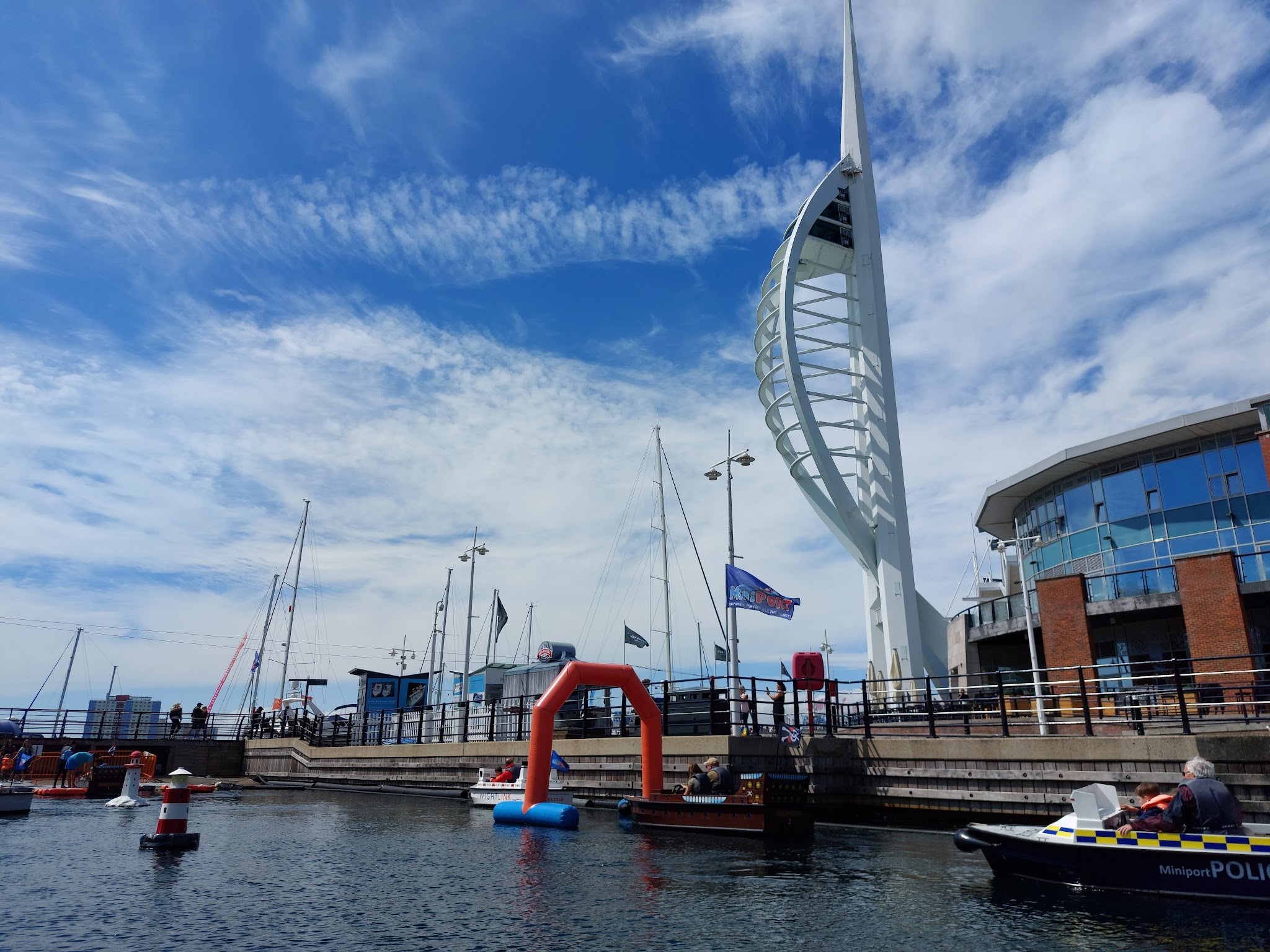 Hafenbecken mit Segelbooten; Spinnaker Tower ragt in blauen Himmel, kleine Boote und orangefarbener Torbogen im Vordergrund