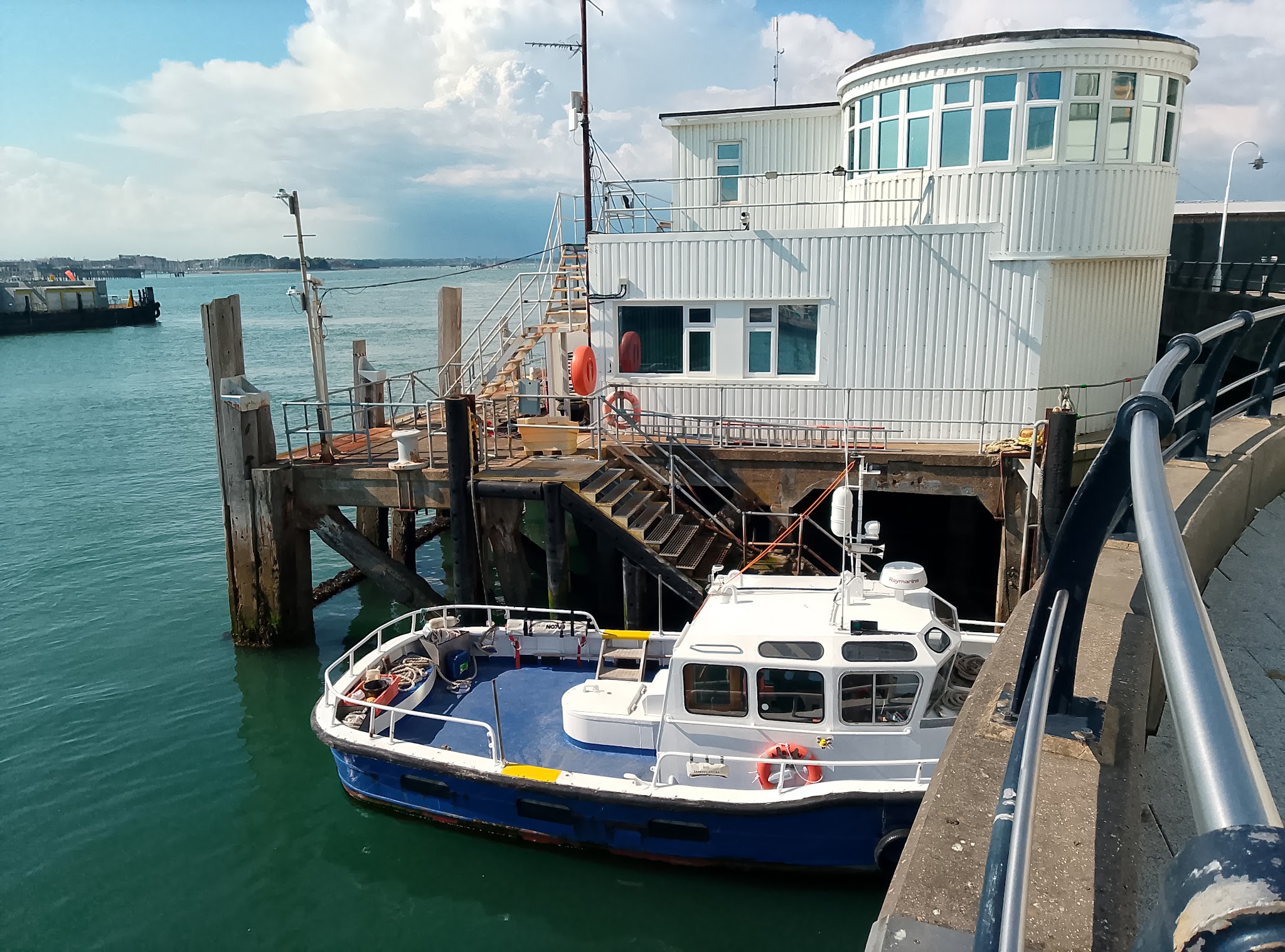 Kleines blau-weißes Boot liegt an hölzernem Anleger vor rundem, weißem Hafenhaus; ruhiges türkisgrünes Meer.