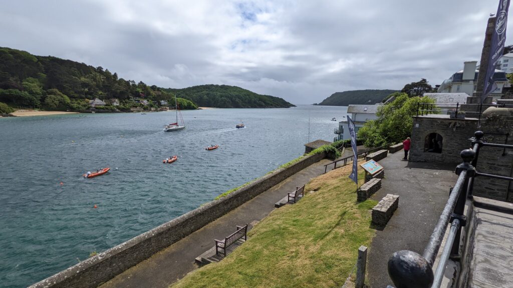 Küstenbucht mit Segelboot und roten Schlauchbooten, bewaldete Hügel gegenüber, Promenadenweg mit Bänken im Vordergrund