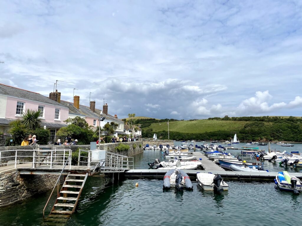 Pastellfarbene Küstenhäuser am Steg, Menschen sitzen im Café; davor viele kleine Boote, grüne Hügel dahinter.