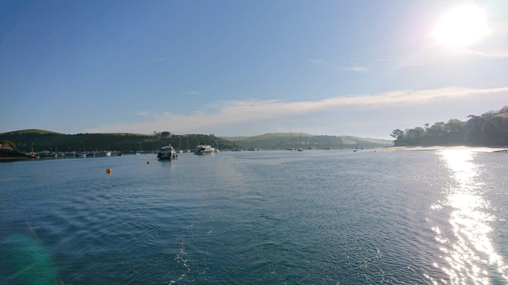 Ruhiger Küstenhafen: Segelboote ankern vor grünen Hügeln, tief stehende Sonne spiegelt hell im Wasser.