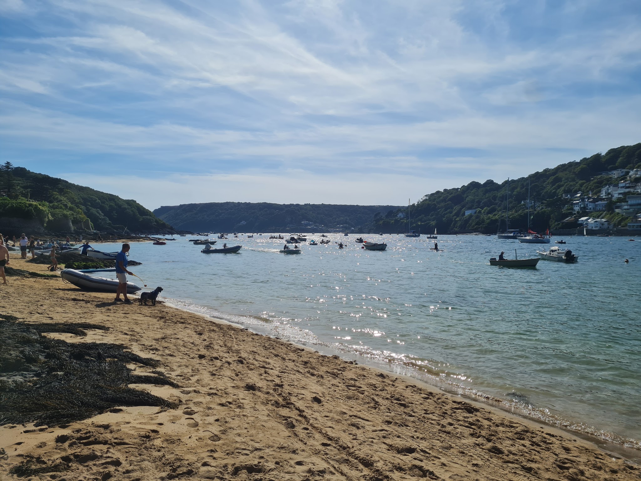 Sandy beach with people and a dog; numerous small boats float on a sun-sparkled bay framed by tree-covered hills.