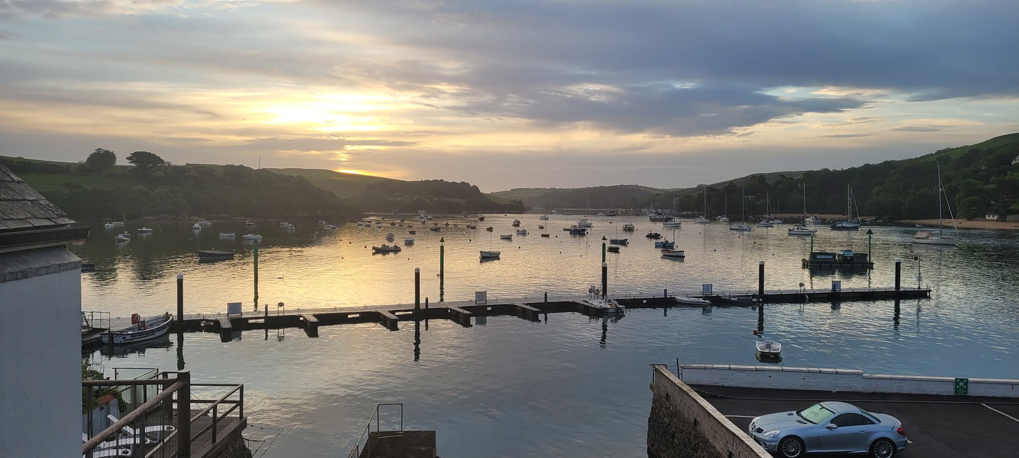 Sonnenuntergang über ruhiger Flussmündung; viele Boote an Stegen, goldenes Licht spiegelt sich im Wasser.