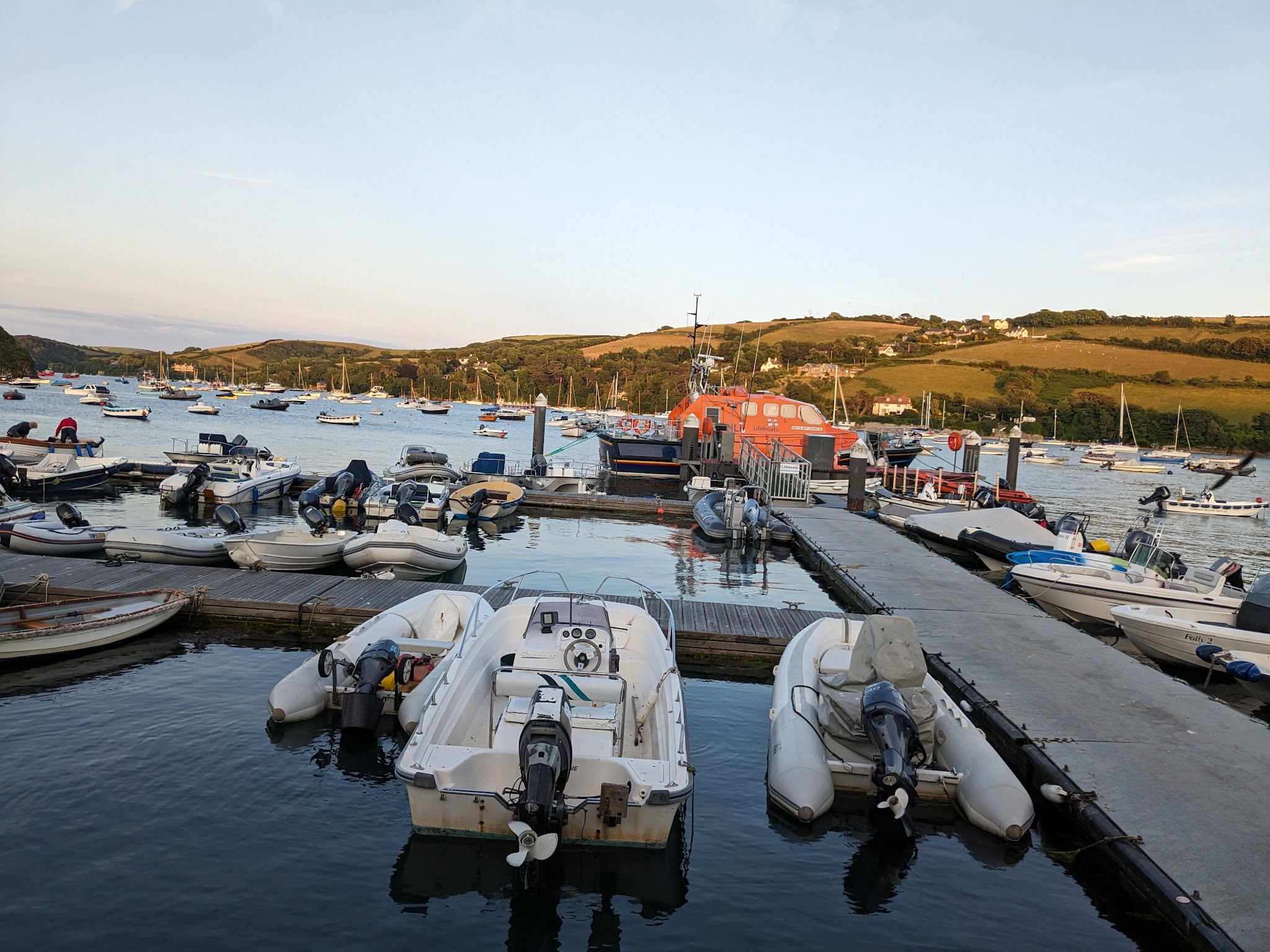 Hafen im Abendlicht: mehrere Schlauchboote an Stegen, zentrales orange Rettungsboot, ruhige Bucht vor grünen Hügeln