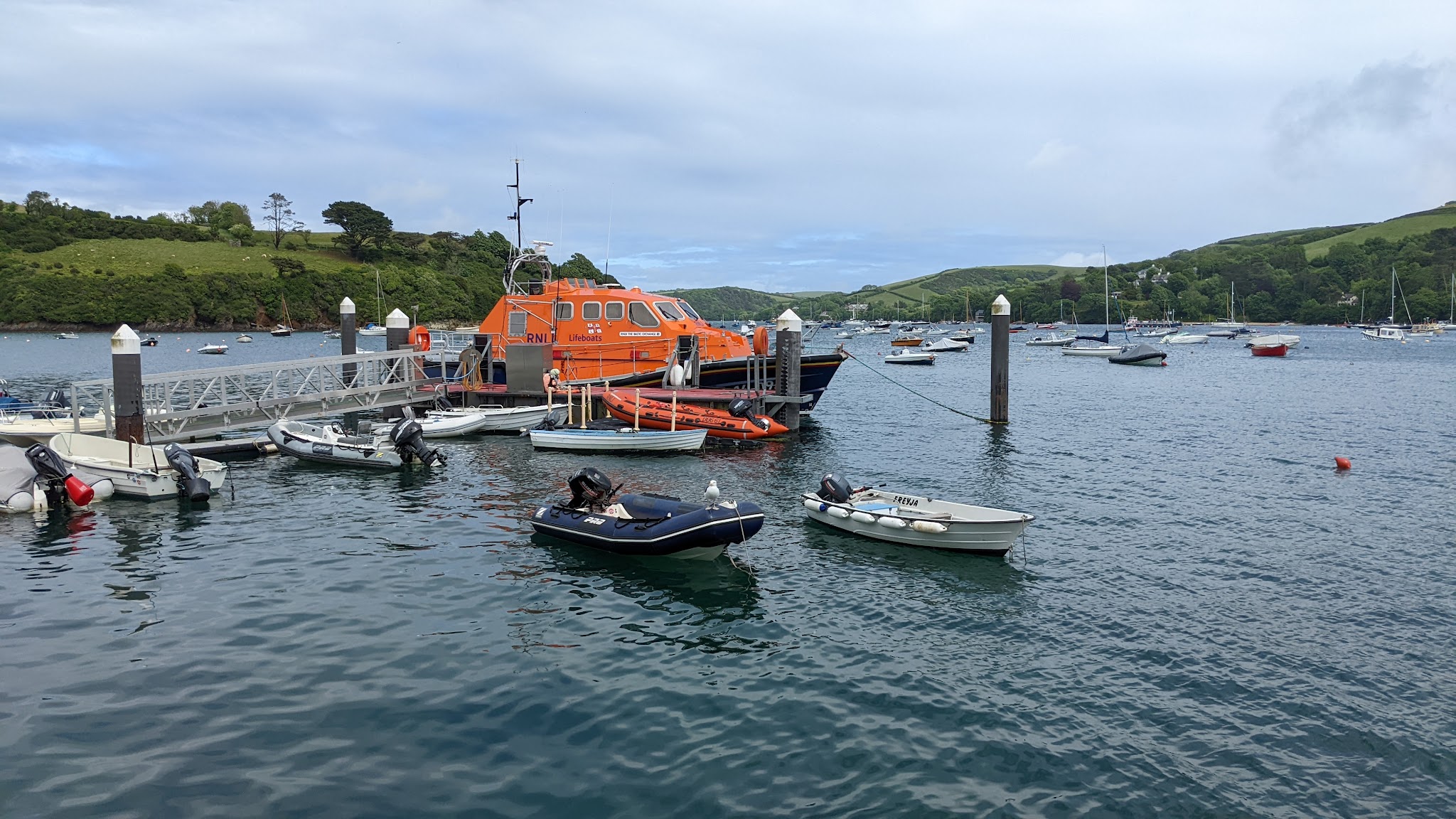 Knallorange RNLI-Rettungsboot am Steg; ringsum Dinghys und Segelboote vor grünen Hügeln in ruhiger Bucht.