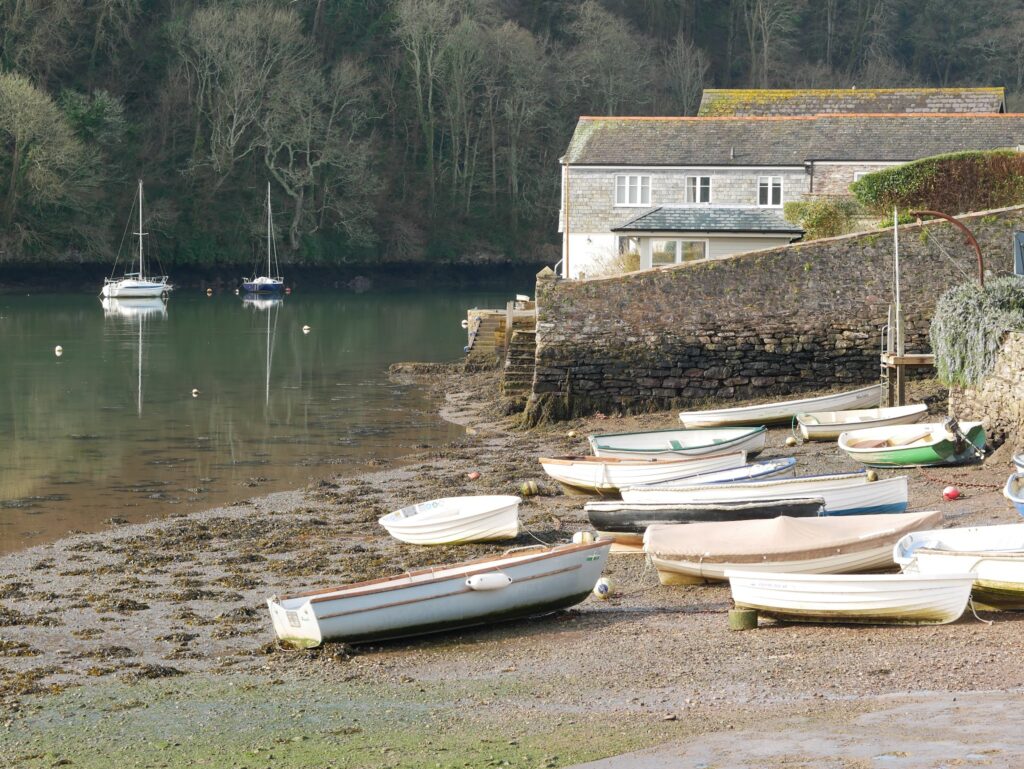 Bei Ebbe ruhen Ruderboote auf dem schlammigen Flussufer, zwei Segelboote spiegeln sich im stillen Wasser.