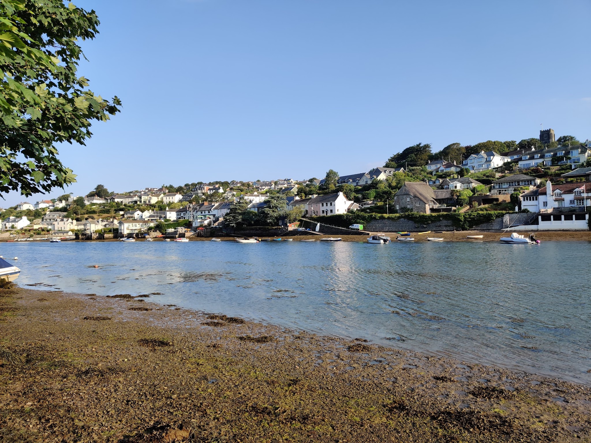 Küstenort mit bunten Hanghäusern, Boote auf ruhigem Wasser bei Ebbe, Laubzweig links im Vordergrund