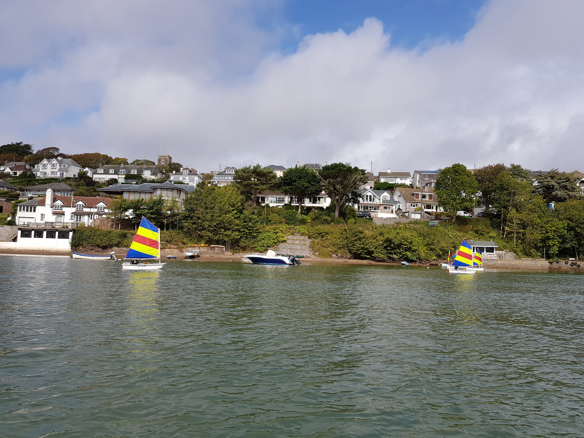 Zwei Segeljollen mit gelb-blau-rot gestreiften Segeln gleiten über ruhige Bucht; dahinter weiße Häuser am grünen Hang.