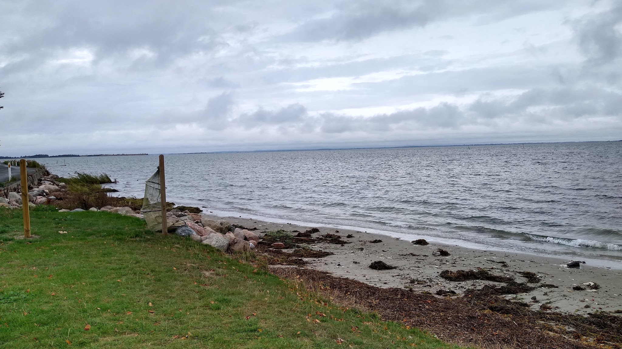 Bewölkter Himmel über ruhiger Ostseeküste, kleine Wellen treffen auf schmalen Sandstrand neben grünem Gras und Steinen.