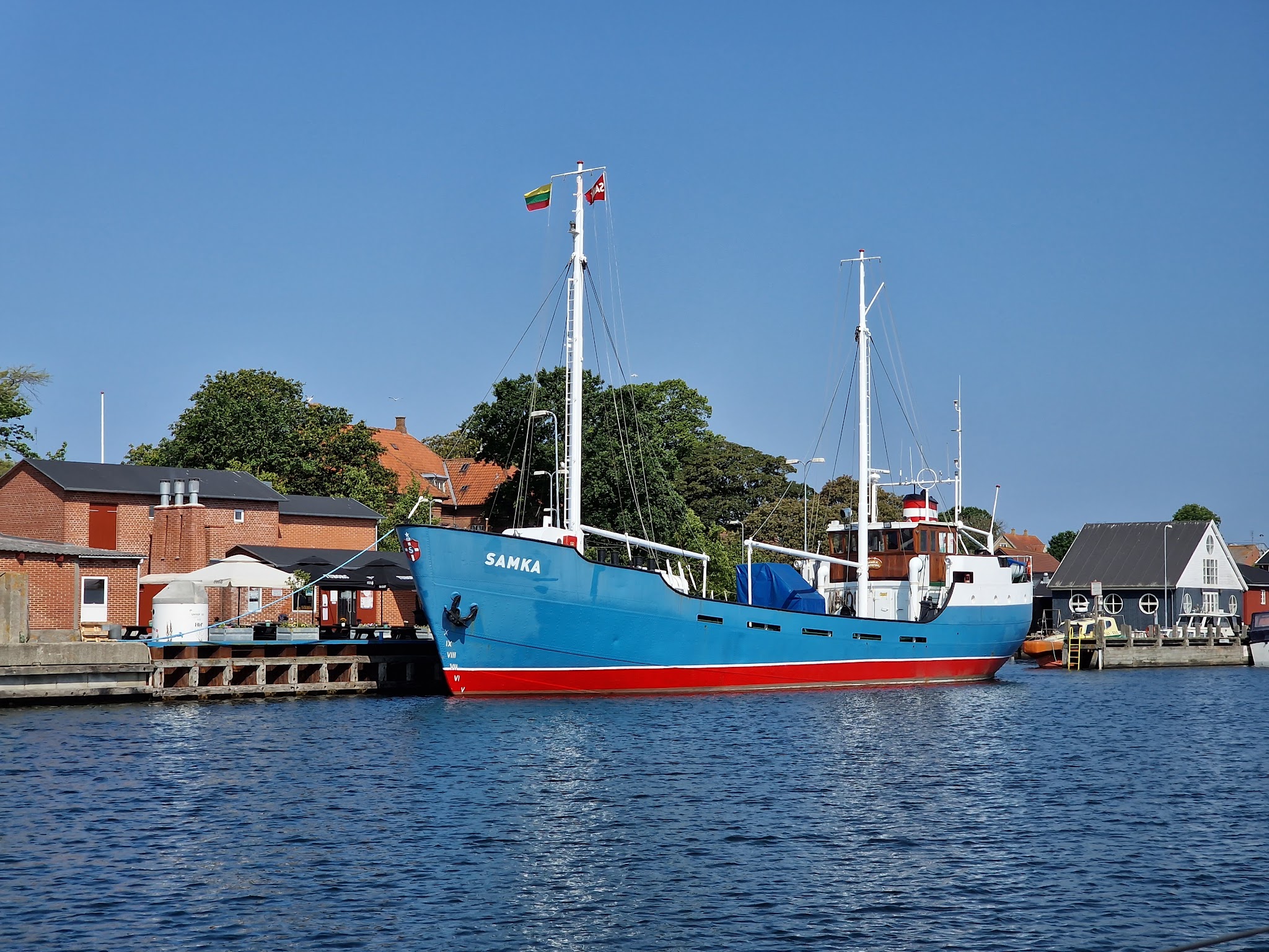 Blau-rotes Schiff „SAMKA“ liegt am Kai vor Backsteinhäusern unter klarem Himmel.