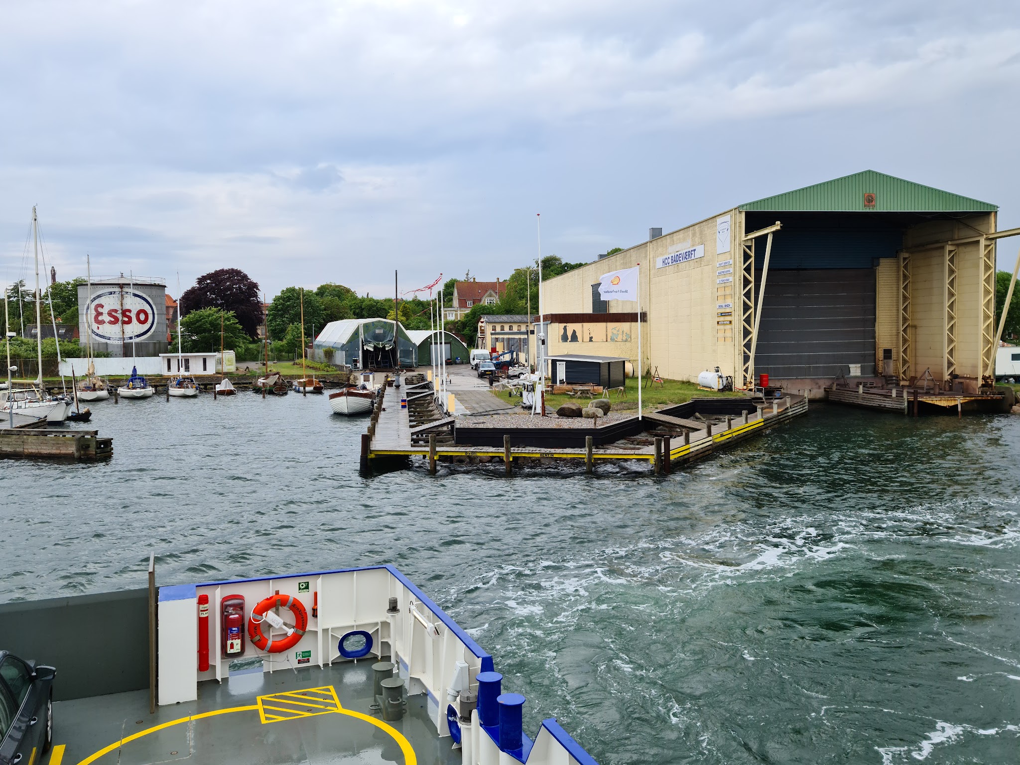 Fährdeck verlässt kleinen Hafen mit Segelbooten; rechts große Werfthalle, links runder ESSO-Tank.
