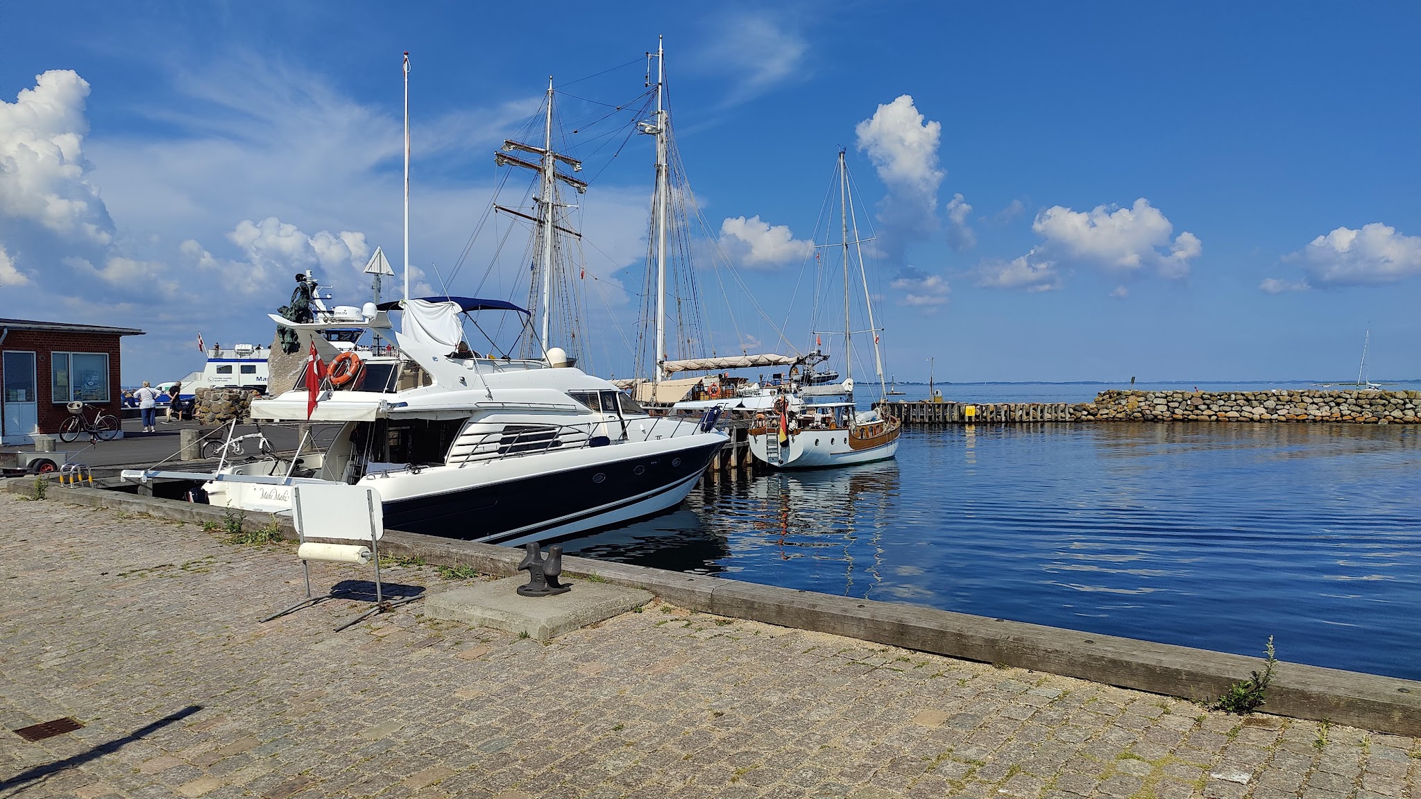 Motorjacht und mehrere Segelboote liegen in ruhigem Hafen; blauer Himmel, vereinzelte Wolken.