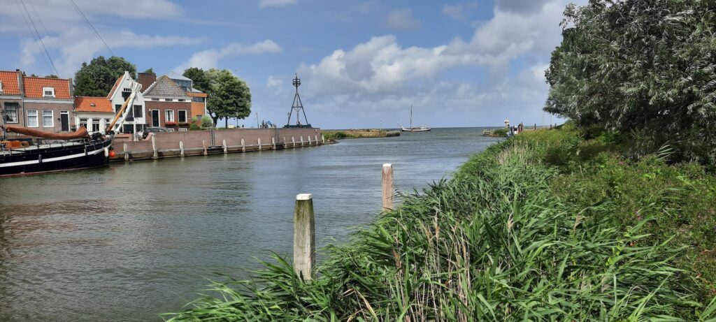 Kleiner Hafenkanal; links Reihenhäuser und Segelboot, rechts Schilf und Baum, Wasser mündet unter blauem Himmel ins Meer