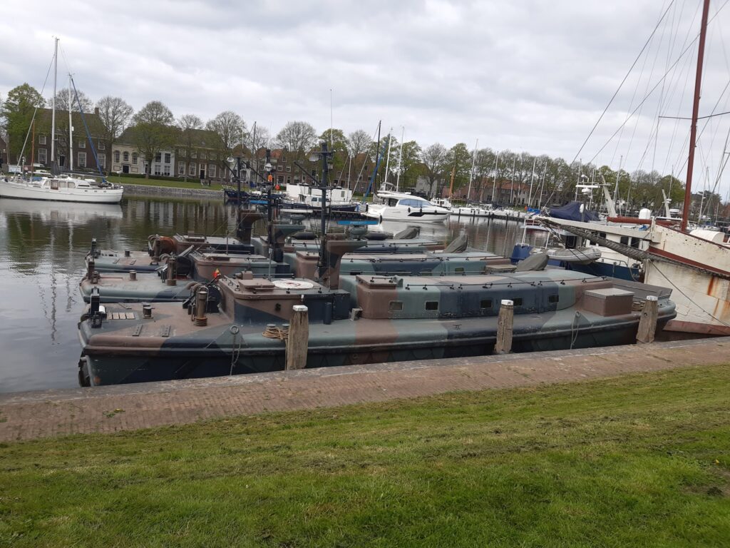 Mehrere tarnfarbene Militärboote liegen im ruhigen Jachthafen; dahinter Segelboote und alte Giebelhäuser.