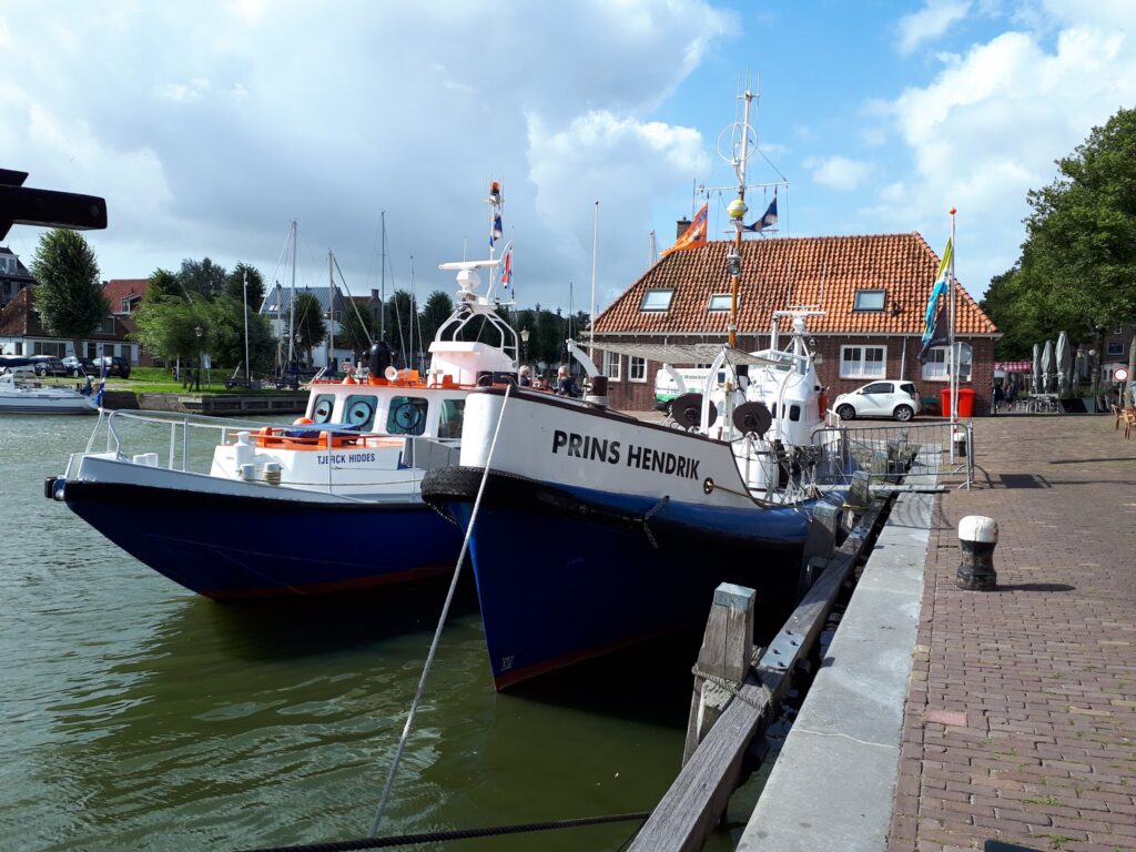 Rettungsboote „Prins Hendrik“ und „Tjerk Hiddes“ vertäut am Kai vor Backsteinhaus; Masten und Wolken im Hintergrund