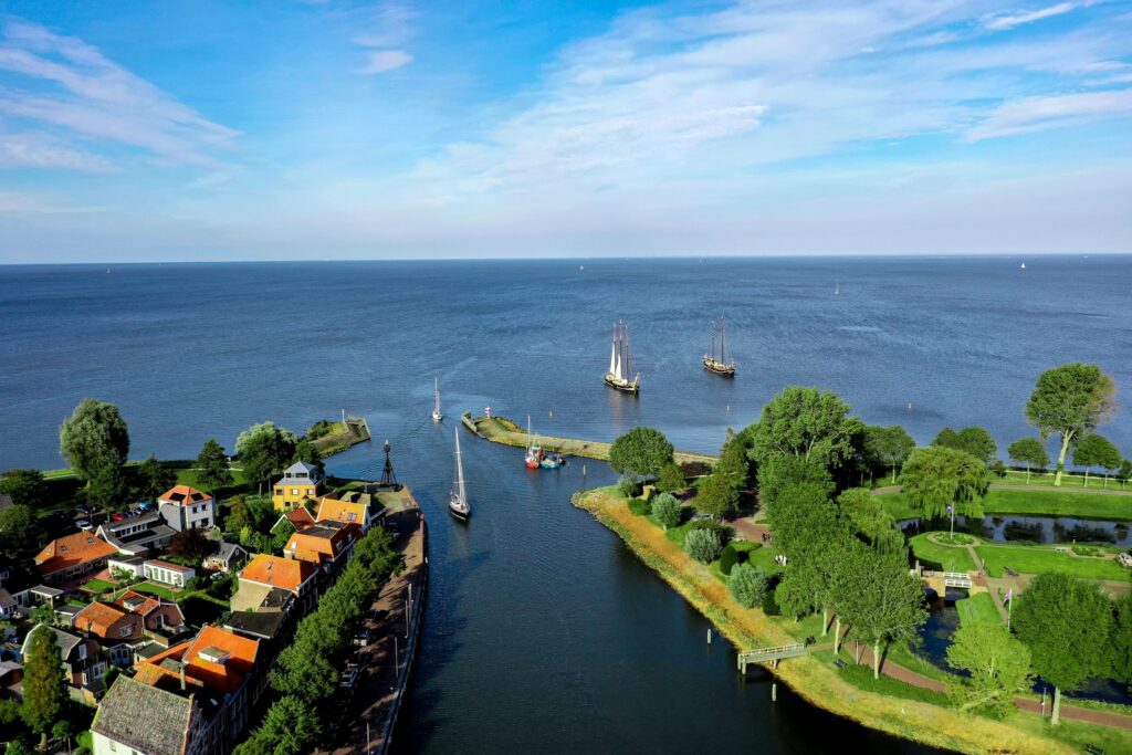 Luftaufnahme: Segelboote fahren durch schmalen Kanal von kleinem Küstendorf mit roten Dächern ins offene Meer.