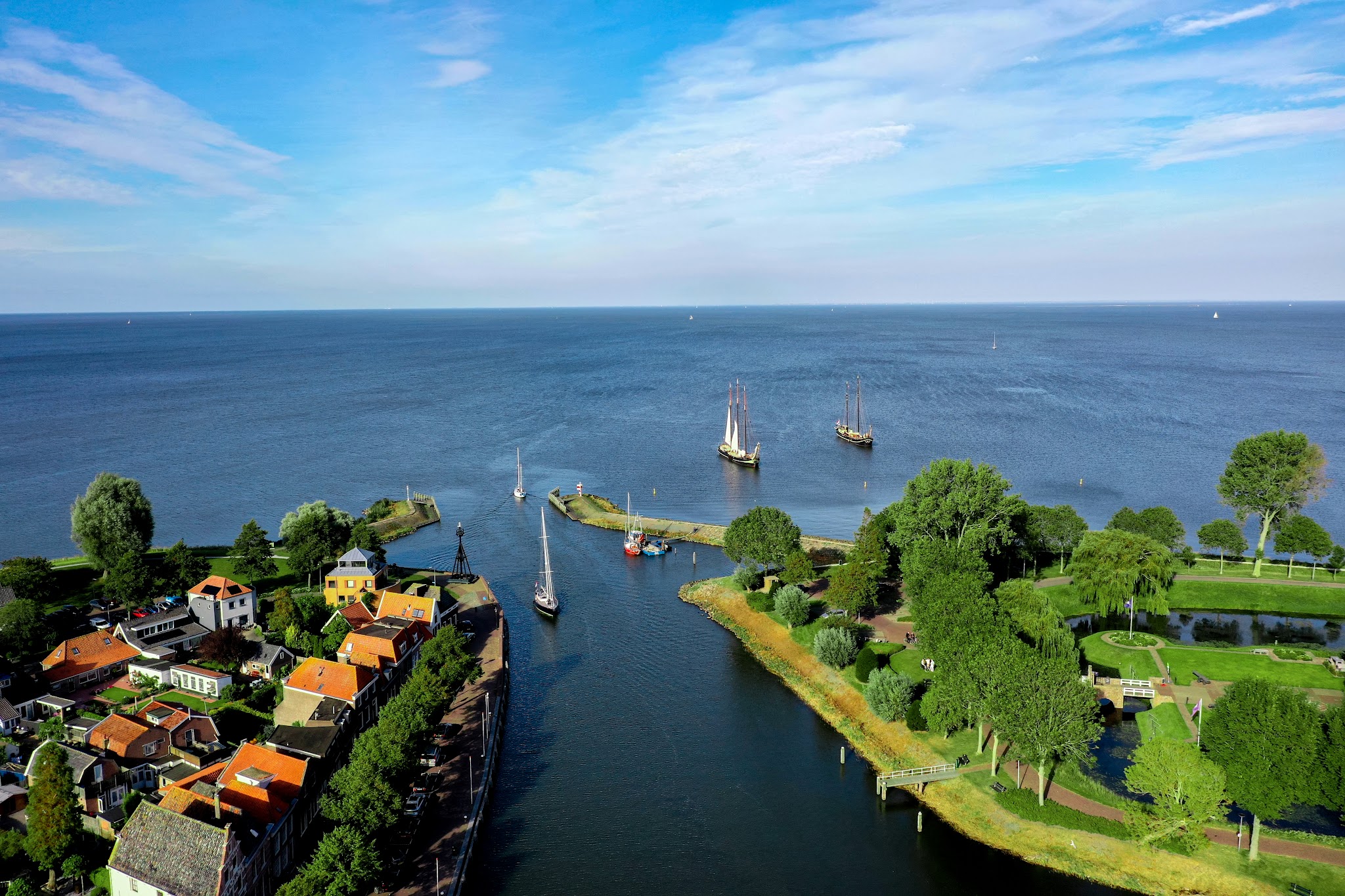 Luftaufnahme: Segelboote fahren durch schmalen Kanal von kleinem Küstendorf mit roten Dächern ins offene Meer.