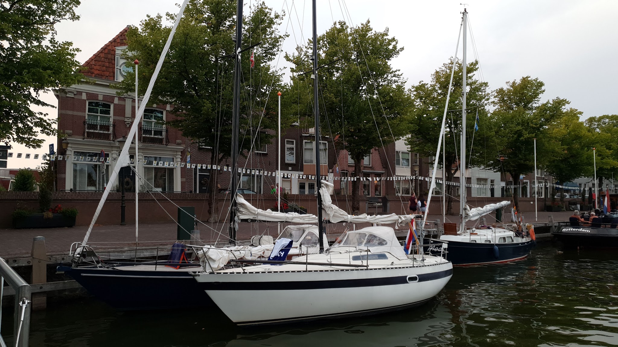 Zwei Segelboote mit gerefften Segeln vertäut in ruhigem Stadthafen vor historischen Backsteinhäusern und Bäumen.