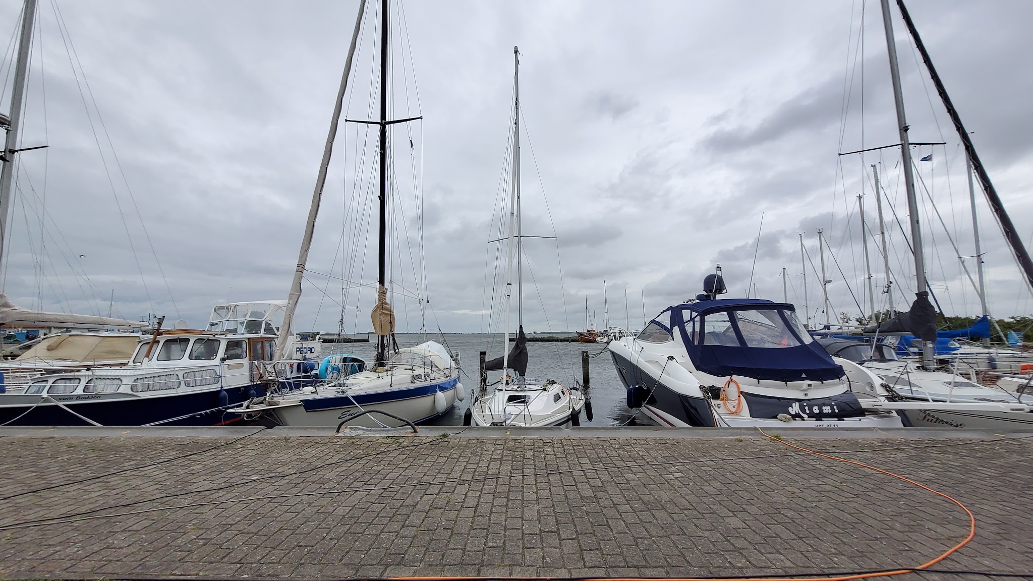 Segel- und Motorboote liegen nebeneinander im ruhigen Hafen, darüber dichter grauer Himmel.
