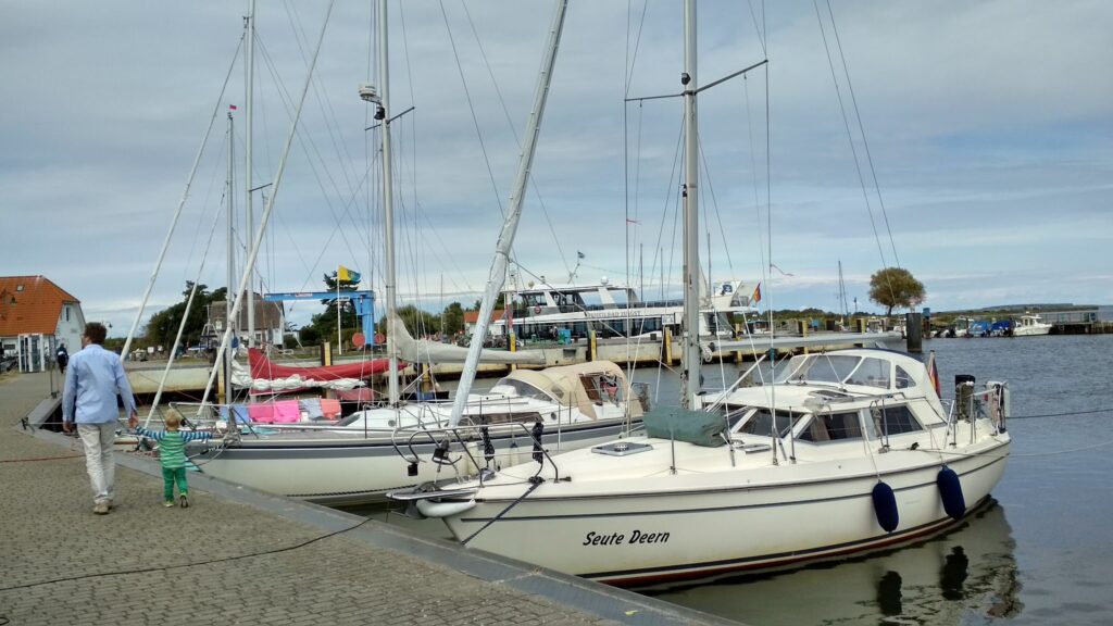 Mann führt kleines Kind an Kajenkante entlang; daneben mehrere festgemachte Segelboote im ruhigen Hafen.
