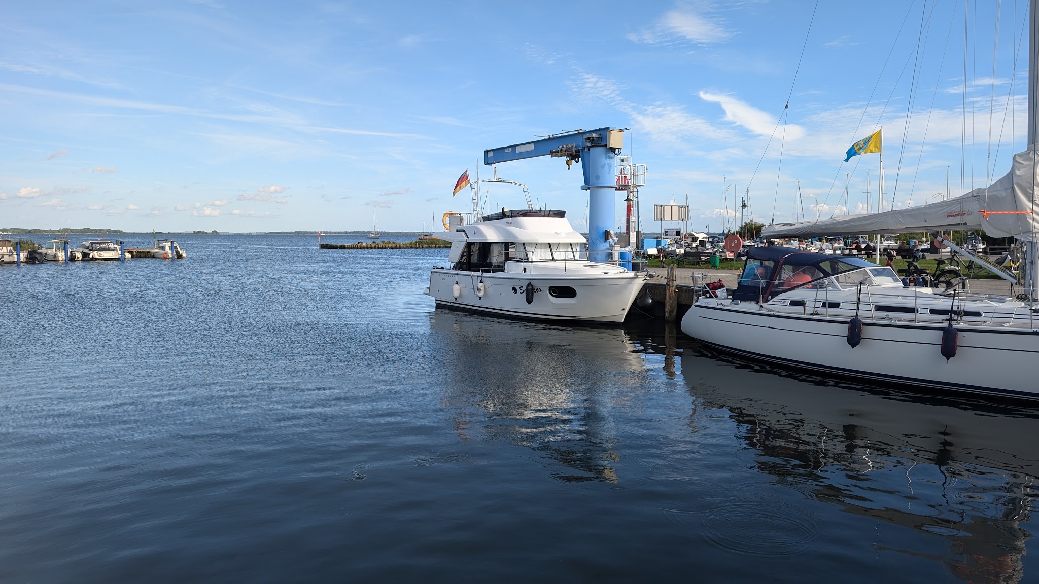 Motor- und Segelboot im ruhigen Hafen, daneben blauer Bootskran; klarer Himmel und Wasser reflektieren.