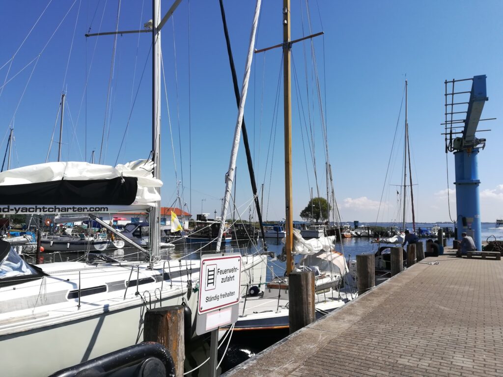 Segelboote im Hafen an sonnigem Steg, blauer Kran und Schild „Feuerwehrzufahrt – ständig freihalten“ im Vordergrund