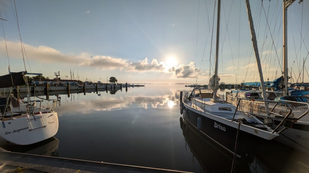 Ruhiger Hafen bei Sonnenaufgang: festgemachte Segelboote spiegeln sich im glatten Wasser unter Wolken