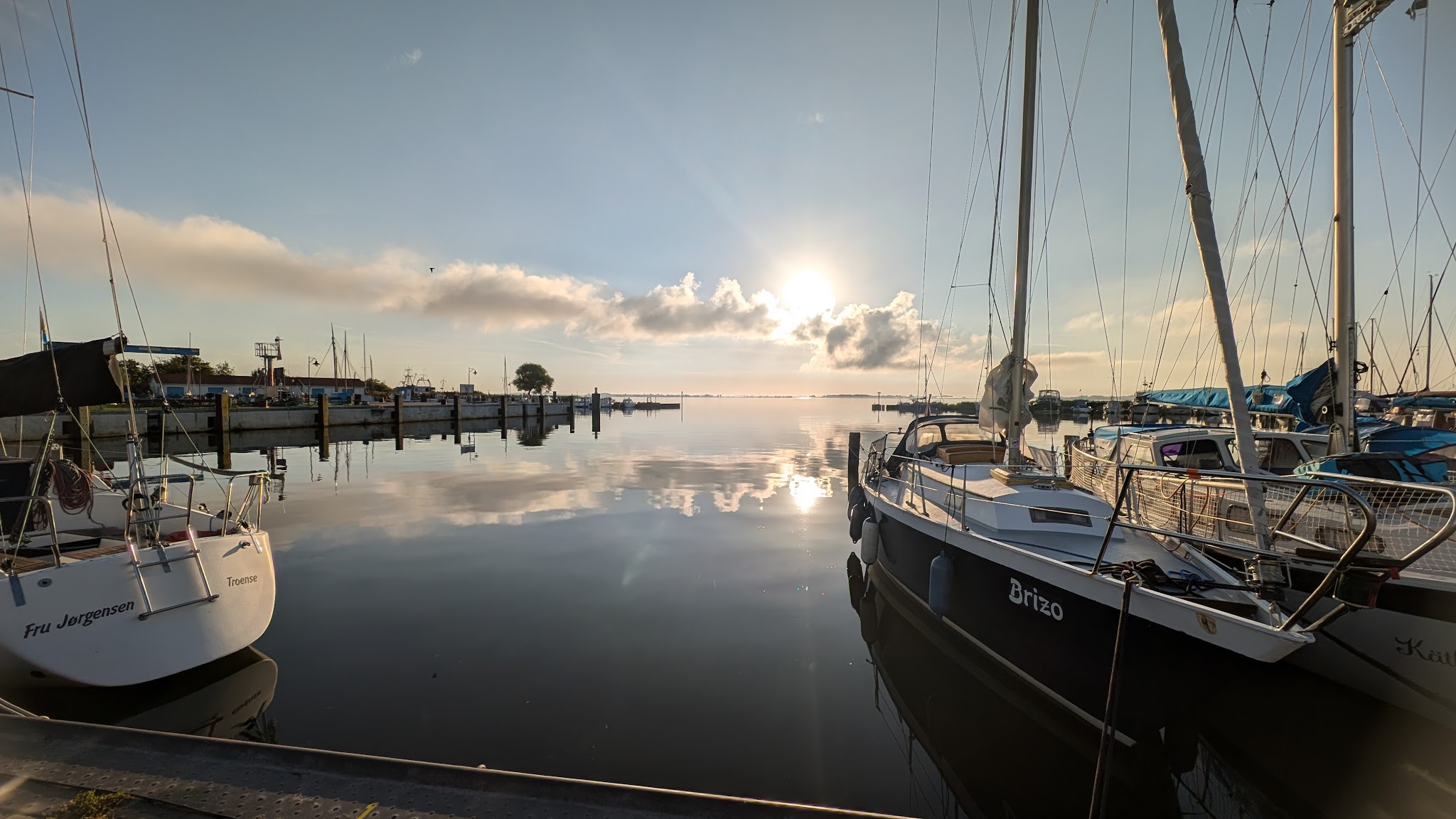 Ruhiger Hafen bei Sonnenaufgang: festgemachte Segelboote spiegeln sich im glatten Wasser unter Wolken