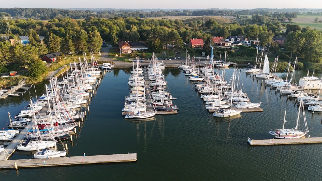 Luftaufnahme eines kleinen Yachthafens mit dicht aneinandergereihten Segelbooten, umgeben von Wald und Dorfhäusern