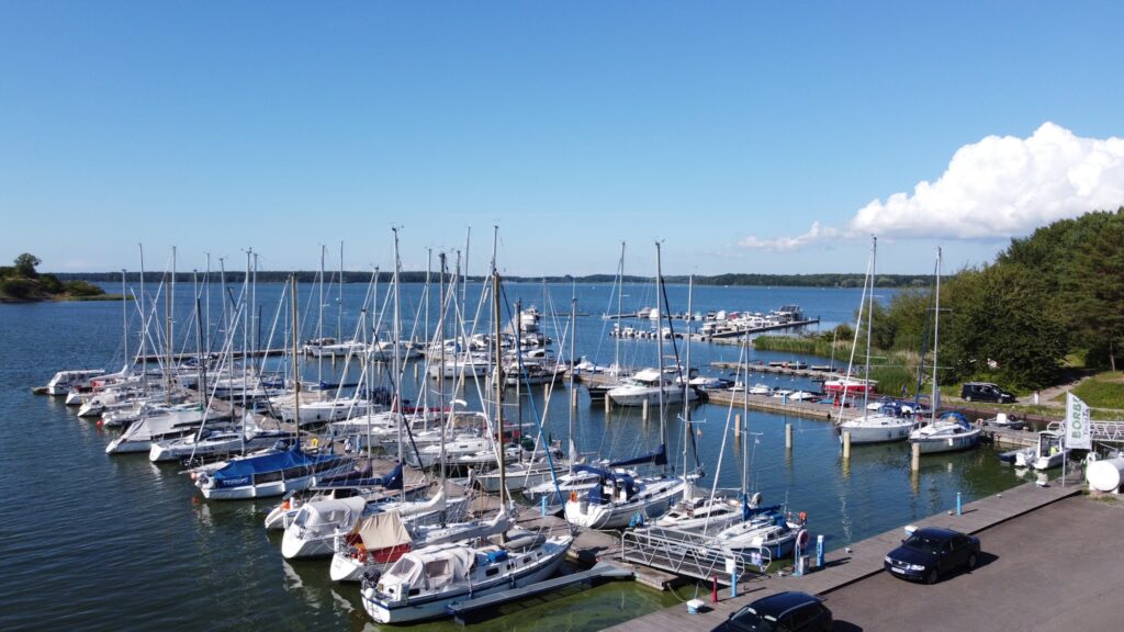Sonniger Hafen mit dicht gereihten Segel- und Motorbooten an Holzstegen; ruhiges blaues Wasser, Wald am Ufer.