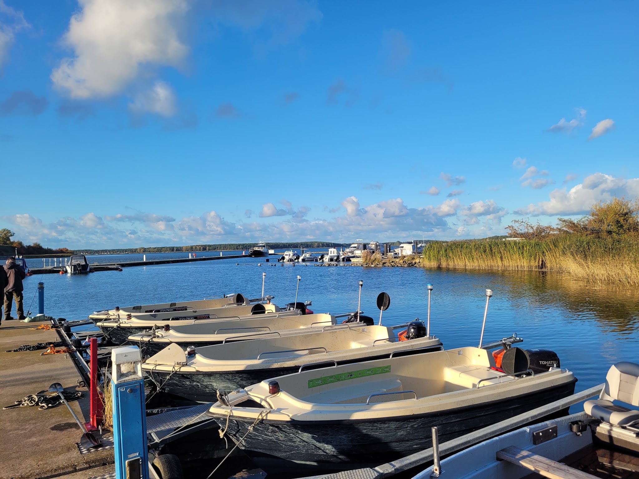 Mehrere kleine Motorboote liegen an einem Steg; ruhiges Wasser, blauer Himmel mit vereinzelten Wolken