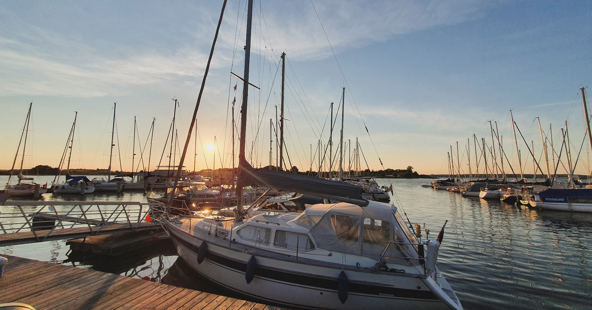 Sonnenuntergang im Yachthafen; zahlreiche Segelboote liegen vertäut, Masten spiegeln sich im ruhigen Wasser.