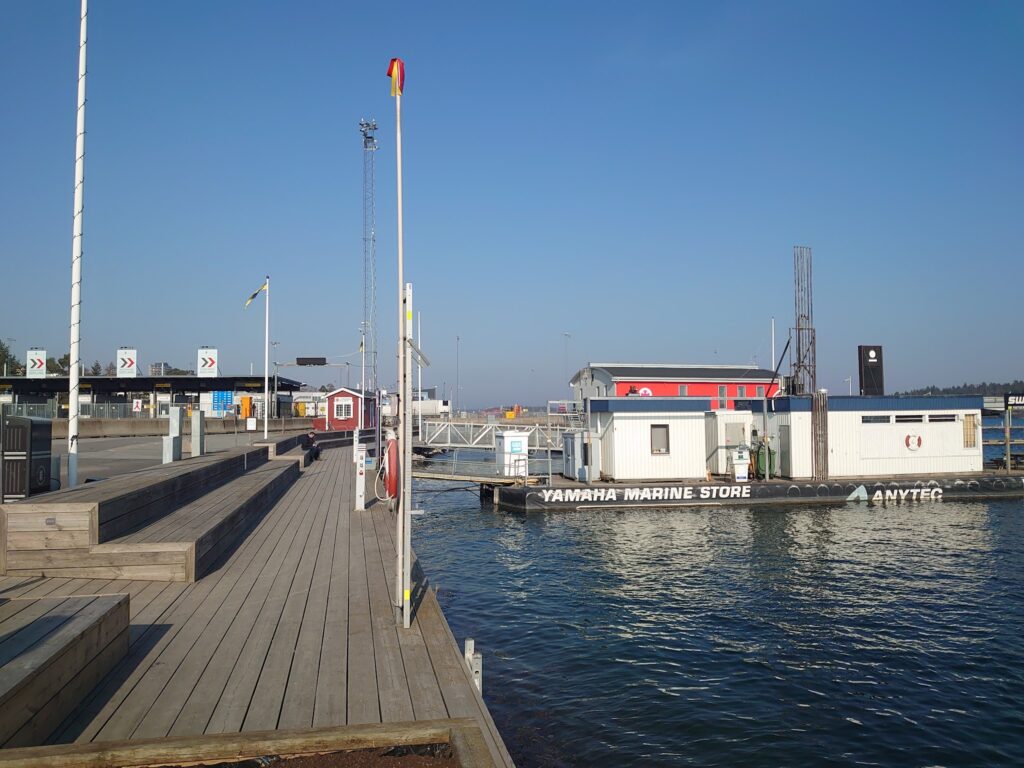 Holzsteg mit Sitzstufen führt zu schwimmender Yamaha-Tankstelle; ruhiges Hafenwasser unter klarem Himmel.