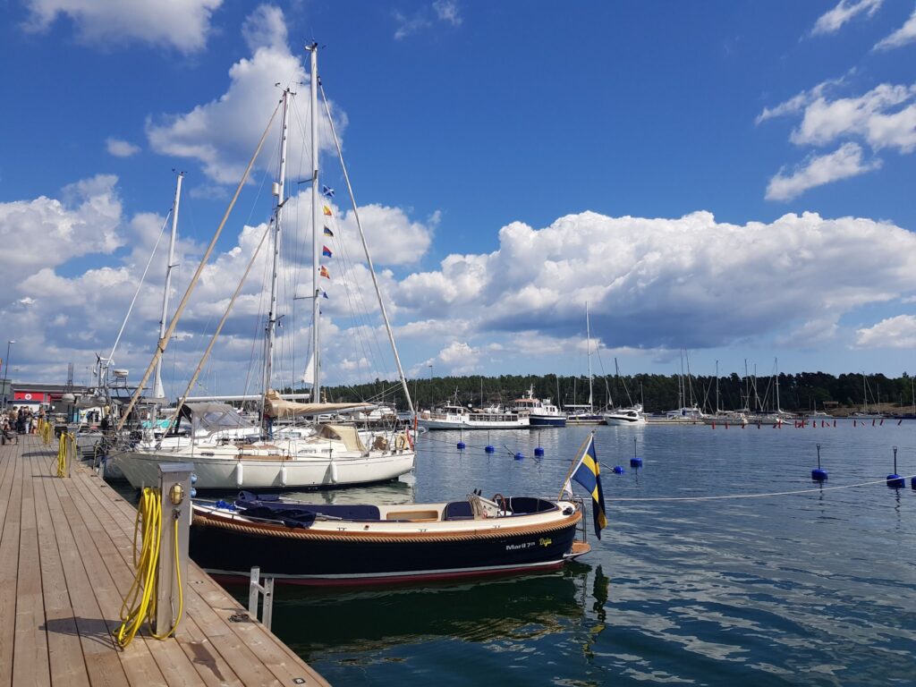 Holzsteg mit Segelbooten im sonnigen Hafen; kleines Boot führt schwedische Flagge, weiße Wolken am Himmel.