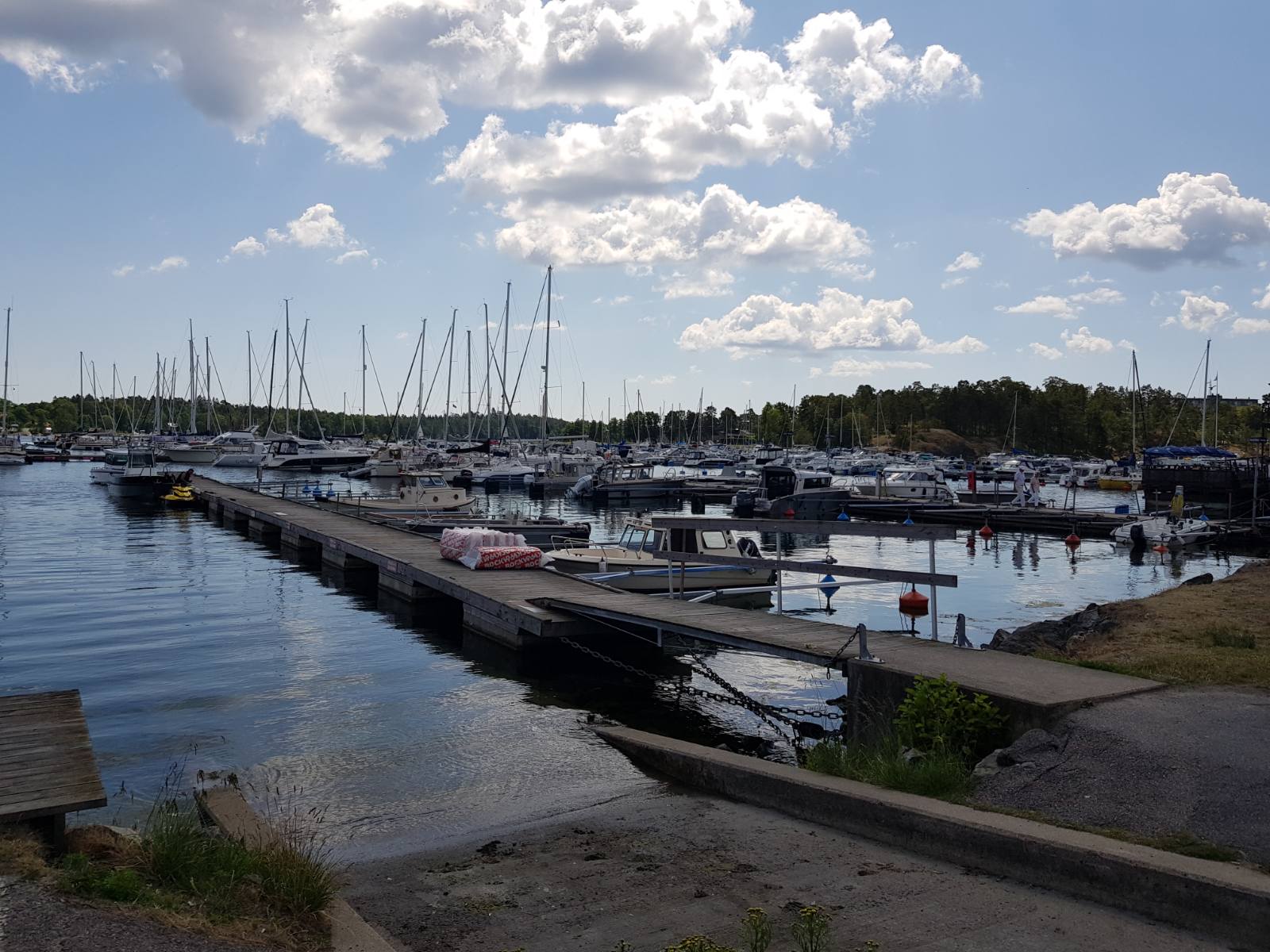 Ruhiger Yachthafen; zahlreiche Segelboote an Stegen, spiegelndes Wasser unter wolkigem Himmel.