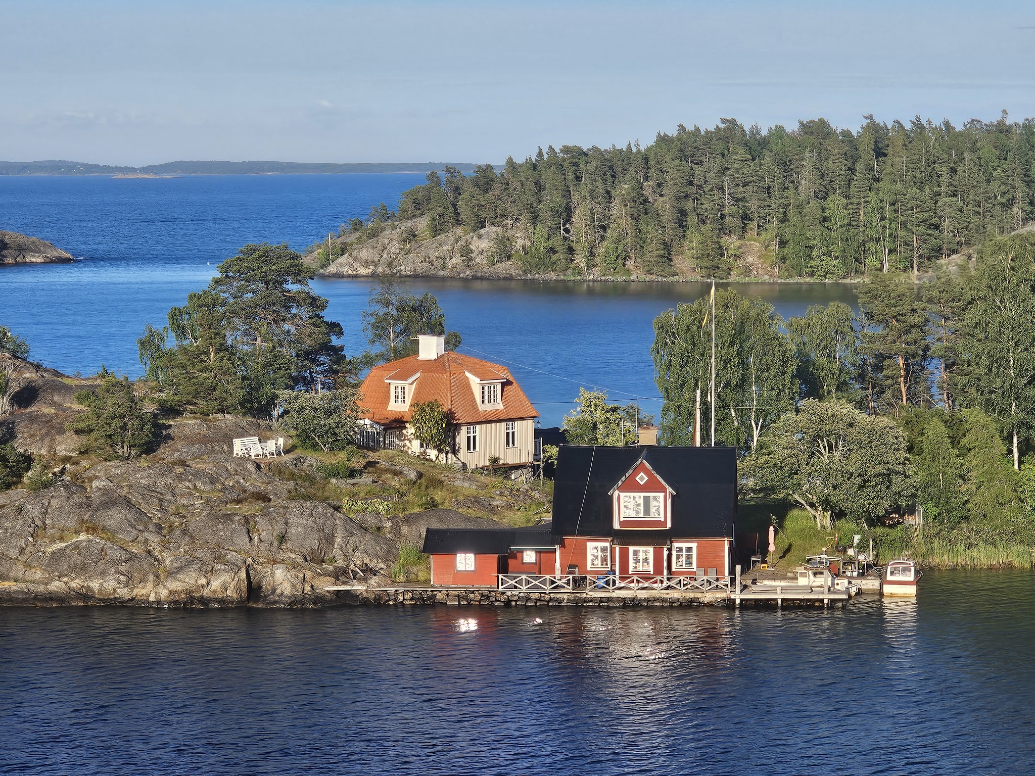 Rotes Holzhaus mit Steg und Boot, daneben beige Haus mit rotem Dach auf felsiger, kiefernreicher Schäreninsel im blauen Meer.