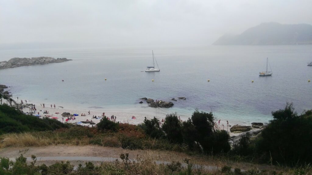 Nebelverhangene Bucht mit weißem Sandstrand, Badenden, Felsen, grüner Böschung und zwei vor Anker liegenden Segelbooten.