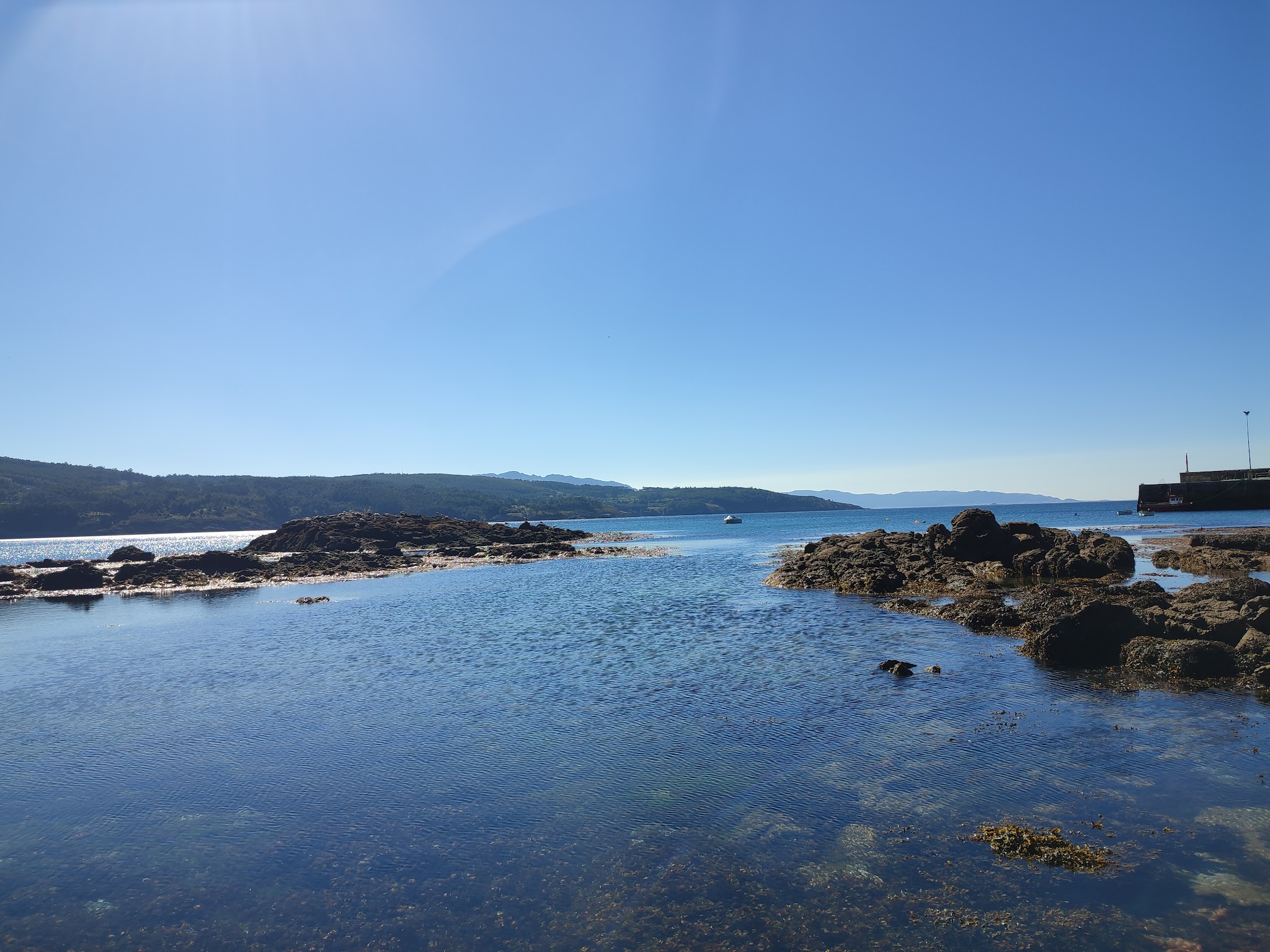 Klares Küstenwasser zwischen dunklen Felsen, kleiner Pier rechts, ferne Hügel und Boot unterm strahlend blauen Himmel.