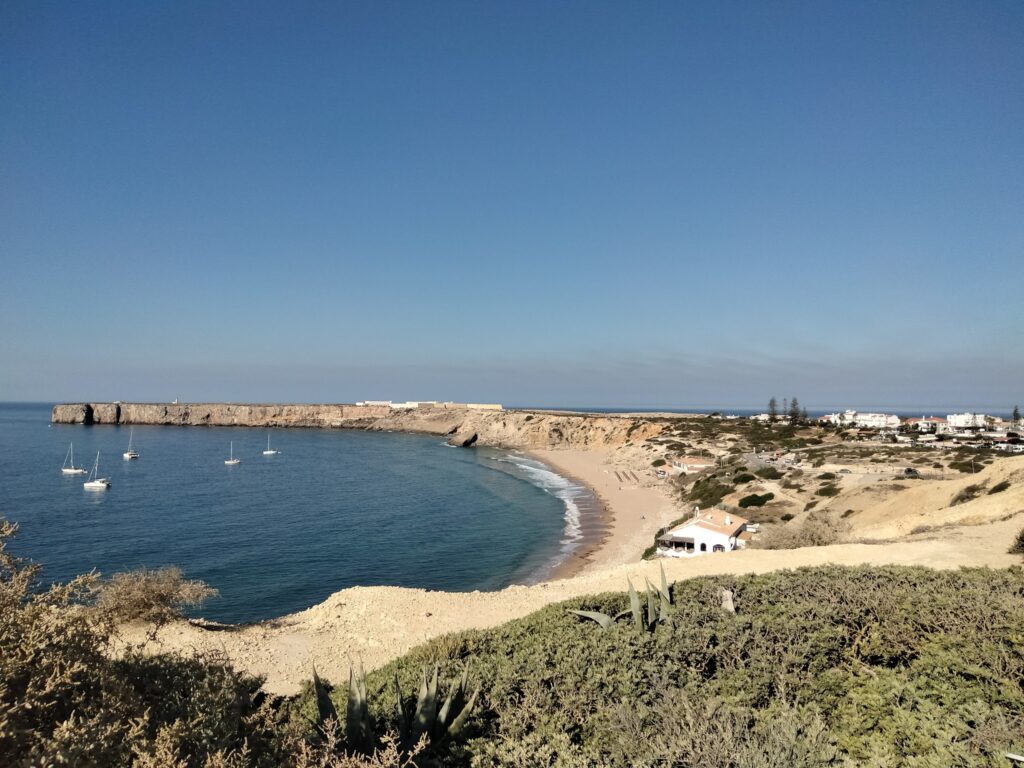 Halbrunde Bucht mit Sandstrand, ruhigem blauem Meer, fünf Segelbooten, steilen Klippen und kleinem Küstenort.