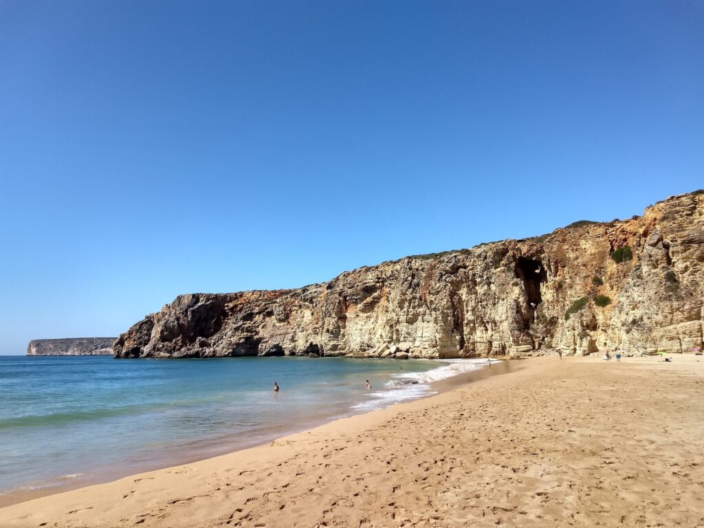 Weite Sandbucht mit türkisblauem Meer vor steilen, hellen Felsklippen unter wolkenlosem Himmel; wenige Badende.