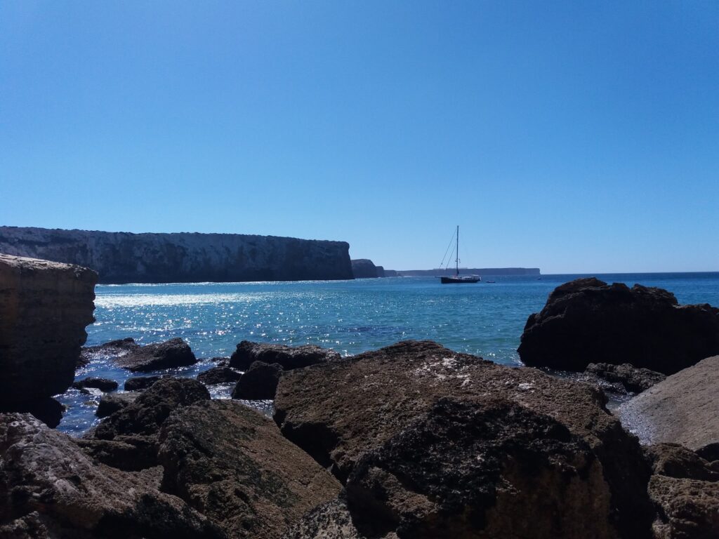 Felsige Küste mit türkisfarbenem Meer, vereinzelte Klippen; ein Segelboot ankert in der sonnigen Ferne.