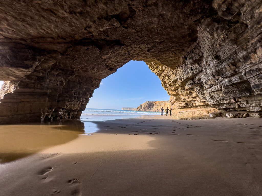 Strandhöhle öffnet sich zum Meer; drei Surfer mit Brettern stehen im Sonnenlicht am Wasserrand, Fußspuren im Sand.