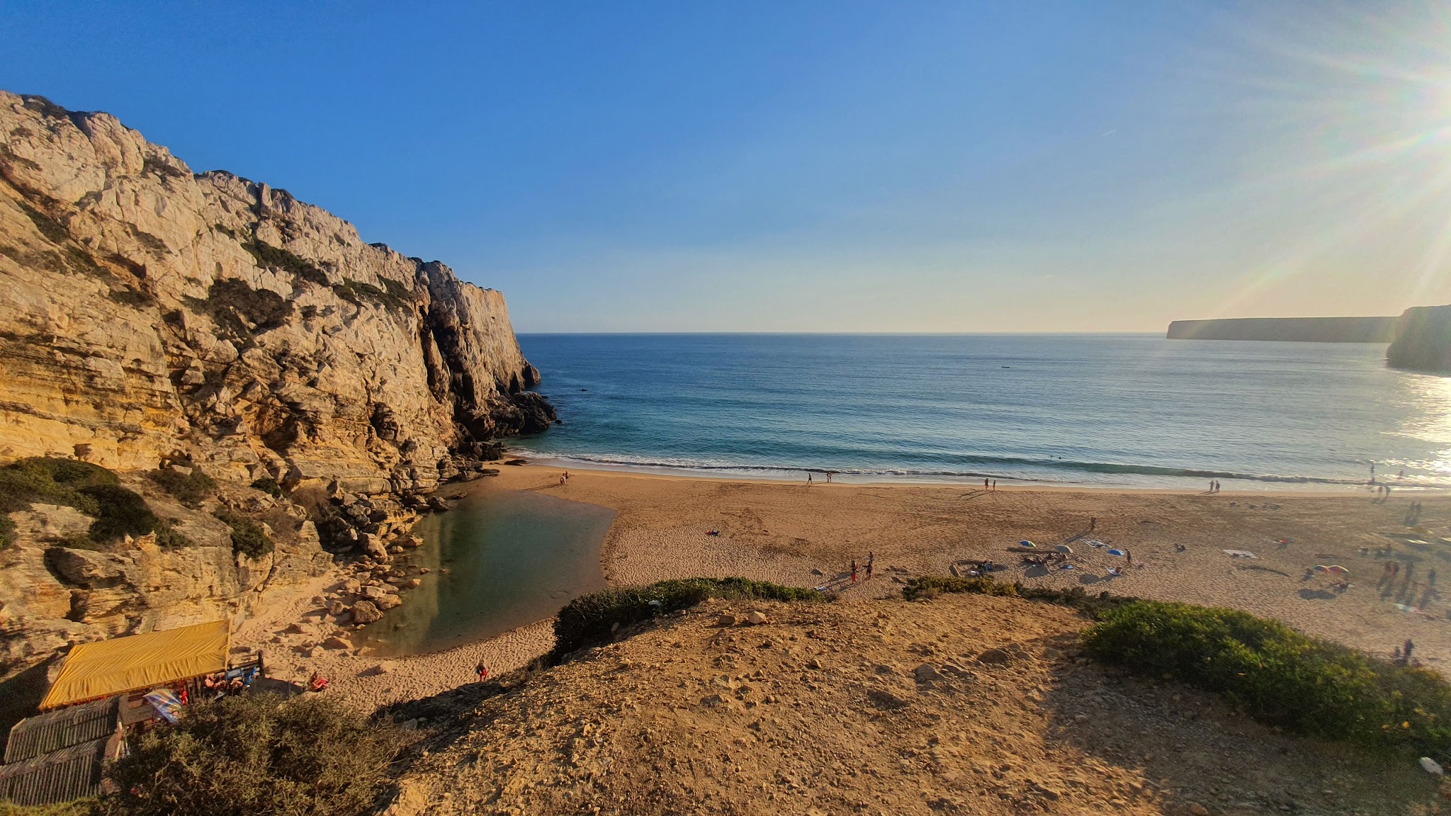Sonniger Sandstrand mit steiler Felsklippe links, ruhigem Meer und wenigen Spaziergängern am Wasser.