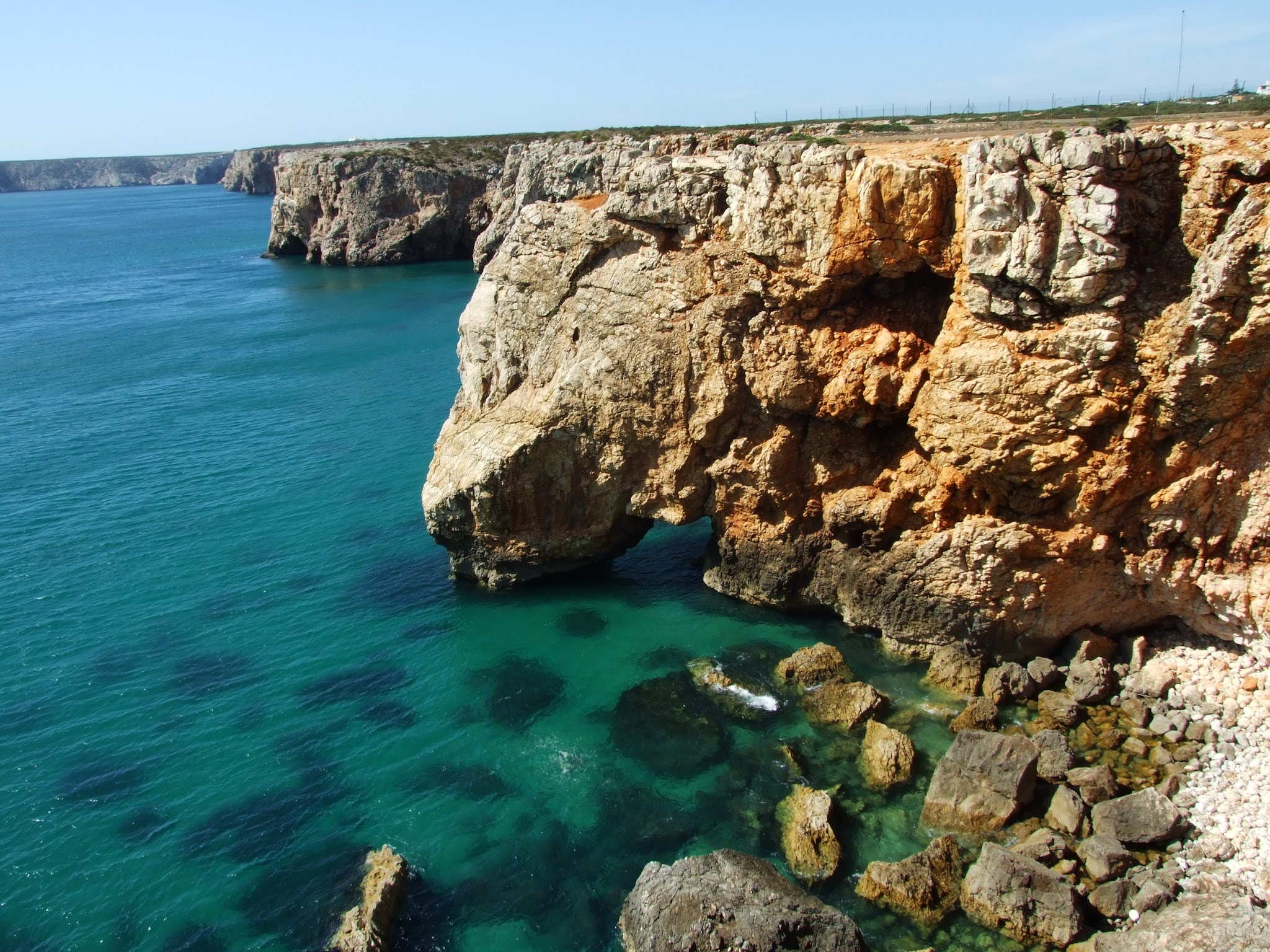 Hohe, zerklüftete Steilküste ragt über türkisblaues Meer; unten im klaren Wasser liegen verstreute Felsen.